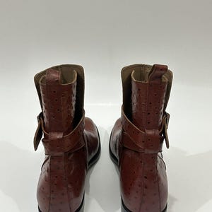 May include: A pair of brown ankle boots featuring a textured, dotted surface pattern. Each boot includes a wrap-around strap with a buckle detail at the side, a pull tab at the top of the shaft, and a solid black sole.