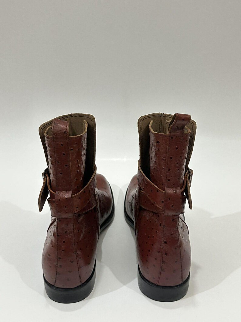 May include: A pair of brown ankle boots featuring a textured, dotted surface pattern. Each boot includes a wrap-around strap with a buckle detail at the side, a pull tab at the top of the shaft, and a solid black sole.