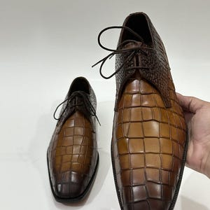 May include: A pair of brown leather dress shoes featuring a prominent crocodile-embossed texture. The shoes have a square toe design, dark brown laces, and a gradient color finish that transitions from a lighter tan to a deep dark brown.