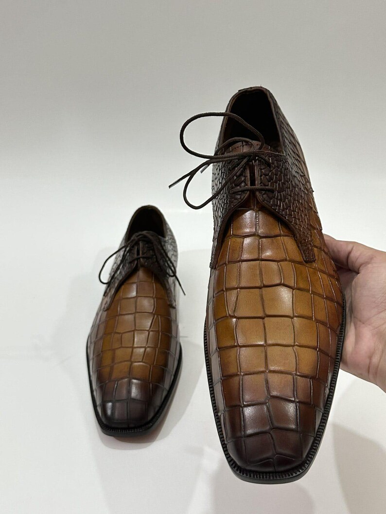 May include: A pair of brown leather dress shoes featuring a prominent crocodile-embossed texture. The shoes have a square toe design, dark brown laces, and a gradient color finish that transitions from a lighter tan to a deep dark brown.