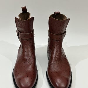 May include: A pair of brown ankle boots featuring a textured ostrich-style leather finish, a rounded toe, a pull tab at the back, and a decorative strap with a metal buckle around the upper shaft, set on a black sole.