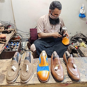 May include: A collection of five leather shoes displayed on a workbench. Two are tan suede loafers with tassels, one is a blue and tan leather brogue, and two are smooth tan leather loafers with tassels. Each shoe features a distinct design.