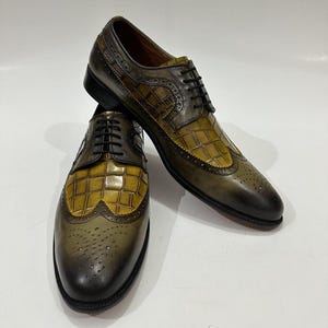 Puede incluir: Un par de zapatos oxford de cuero en color verde oliva y amarillo con dise&ntilde;o de punta de ala, textura de piel de cocodrilo, cordones negros y perforaciones decorativas tipo brogue a lo largo de las costuras y la puntera.