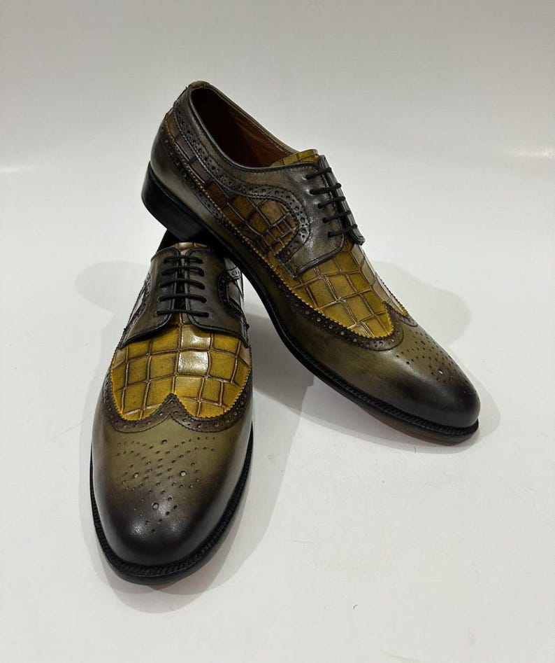 Puede incluir: Un par de zapatos oxford de cuero en color verde oliva y amarillo con dise&ntilde;o de punta de ala, textura de piel de cocodrilo, cordones negros y perforaciones decorativas tipo brogue a lo largo de las costuras y la puntera.