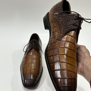 May include: A pair of brown leather dress shoes featuring a distinct embossed crocodile skin texture. The shoes have a lace-up closure, a dark brown heel, and a gradient color finish that transitions from a lighter tan to a deep dark brown.