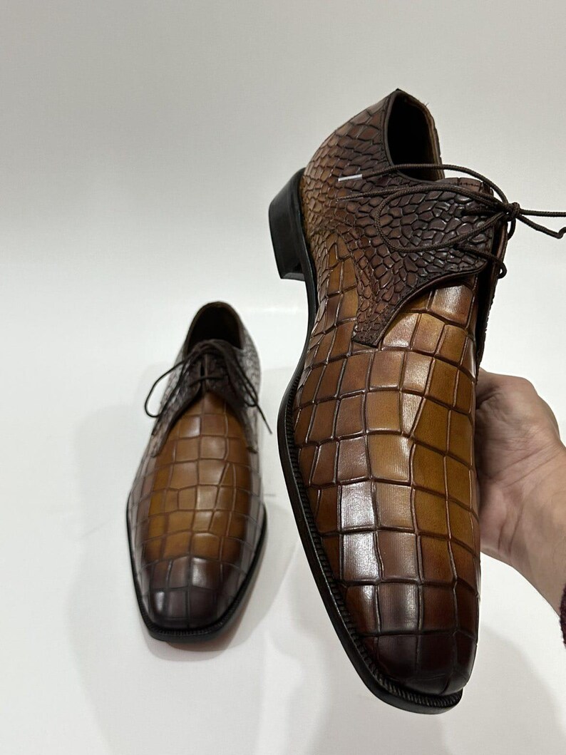 May include: A pair of brown leather dress shoes featuring a distinct embossed crocodile skin texture. The shoes have a lace-up closure, a dark brown heel, and a gradient color finish that transitions from a lighter tan to a deep dark brown.