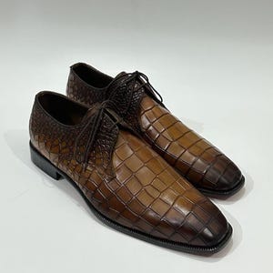 May include: A pair of brown lace-up dress shoes featuring a textured crocodile-embossed pattern. The shoes have a gradient color effect, transitioning from a lighter tan in the center to a darker brown at the edges, with thin brown laces.
