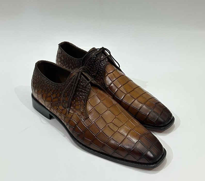 May include: A pair of brown lace-up dress shoes featuring a textured crocodile-embossed pattern. The shoes have a gradient color effect, transitioning from a lighter tan in the center to a darker brown at the edges, with thin brown laces.