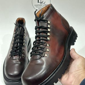 May include: A pair of dark brown leather ankle boots featuring black laces, silver-toned metal D-ring eyelets, and a thick black lugged rubber sole. The boots have a smooth finish and a logo reading LEATHER LORD printed in the background.