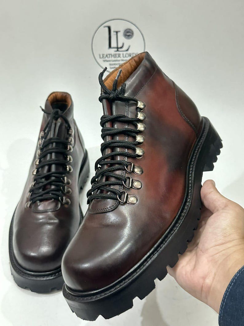 May include: A pair of dark brown leather ankle boots featuring black laces, silver-toned metal D-ring eyelets, and a thick black lugged rubber sole. The boots have a smooth finish and a logo reading LEATHER LORD printed in the background.