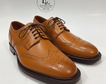 May include: A pair of tan leather wingtip brogue shoes with dark brown laces and a dark brown sole. The shoes feature decorative perforations along the seams and toe cap. A circular logo with the text LEATHER LORDS is visible above the shoes.
