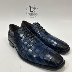 May include: A pair of navy blue leather oxford shoes with a textured crocodile skin pattern, black laces, and a square toe. The shoes feature a gradient finish from dark blue to black at the toe. A logo with the text LEATHER LORDS is visible.