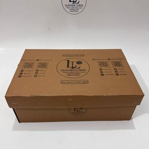 May include: A rectangular brown cardboard box featuring the black Leather Lords logo, text, and QR codes printed on the lid and front side. The branding includes the phrase Where Leather Meets Luxury and lists various leather products.