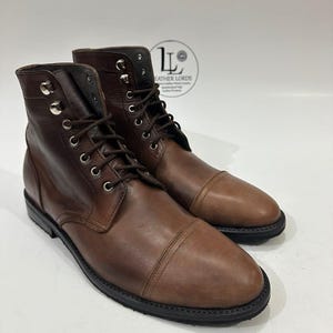 May include: A pair of brown leather lace-up boots featuring a cap toe design, silver-toned metal eyelets, and dark brown laces. The boots have a black sole and a logo stamp on the upper ankle area that reads Leather Lords.