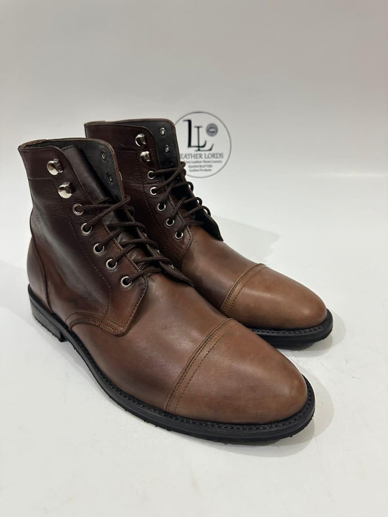 May include: A pair of brown leather lace-up boots featuring a cap toe design, silver-toned metal eyelets, and dark brown laces. The boots have a black sole and a logo stamp on the upper ankle area that reads Leather Lords.
