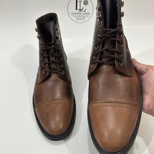 May include: A pair of brown leather cap-toe boots with dark brown laces and silver-toned metal eyelets. The boots feature a smooth leather finish and a black sole. A circular logo with the text Leather Lords is visible in the background.