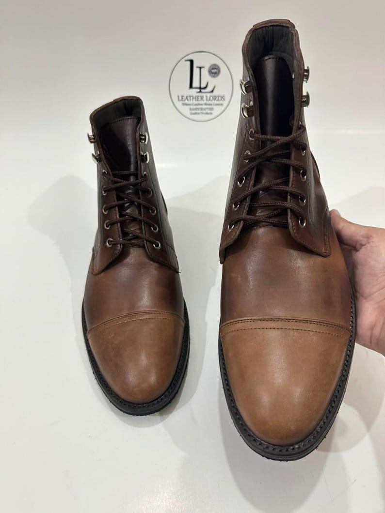 May include: A pair of brown leather cap-toe boots with dark brown laces and silver-toned metal eyelets. The boots feature a smooth leather finish and a black sole. A circular logo with the text Leather Lords is visible in the background.