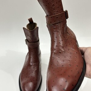 May include: A pair of brown ankle boots featuring a textured ostrich-style leather finish with distinct quill follicles. Each boot includes a side buckle strap, a pull tab at the top of the shaft, and a thick black sole with visible stitching.