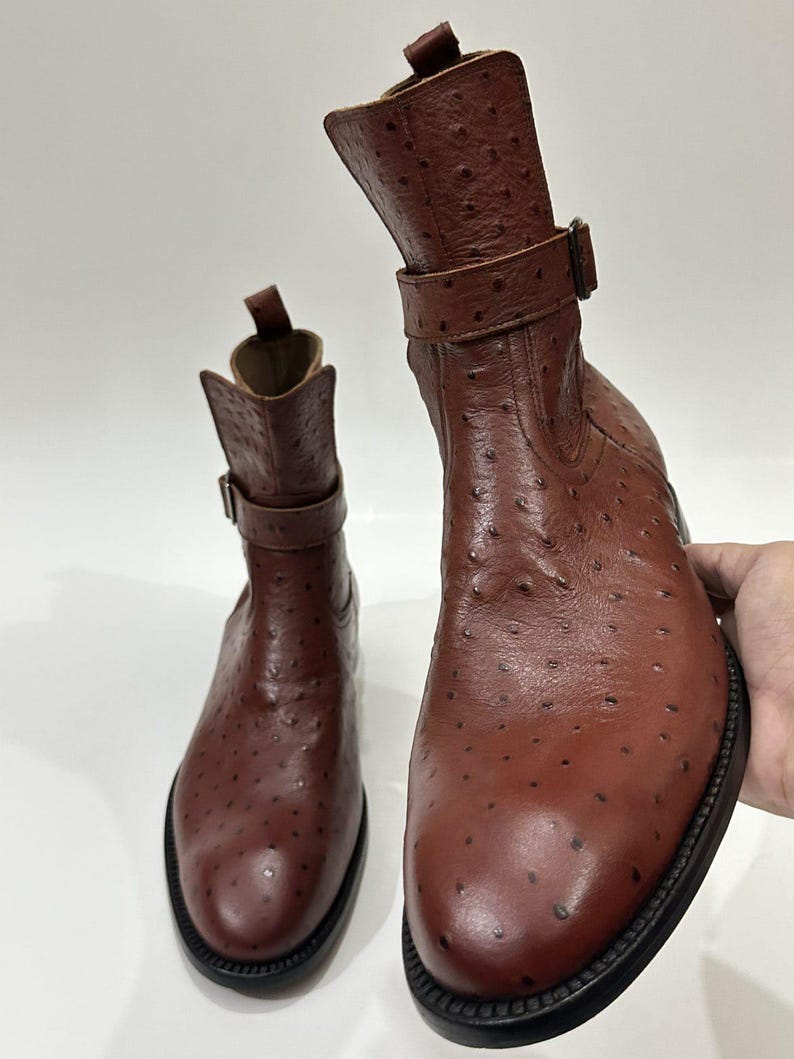 May include: A pair of brown ankle boots featuring a textured ostrich-style leather finish with distinct quill follicles. Each boot includes a side buckle strap, a pull tab at the top of the shaft, and a thick black sole with visible stitching.