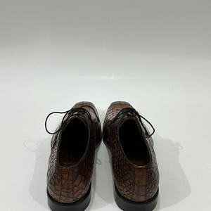 May include: A pair of brown leather dress shoes featuring a textured crocodile skin pattern. The shoes have a lace-up closure with thin dark brown laces, a rounded toe box, and a solid black heel base. The interior lining is a dark brown color.