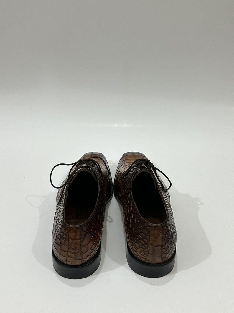 May include: A pair of brown leather dress shoes featuring a textured crocodile skin pattern. The shoes have a lace-up closure with thin dark brown laces, a rounded toe box, and a solid black heel base. The interior lining is a dark brown color.