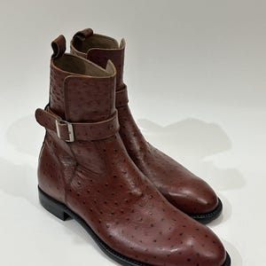 May include: A pair of brown leather ankle boots with a textured surface and a buckle detail. The boots have a rounded toe and a black sole. The boots are approximately 6 inches tall.