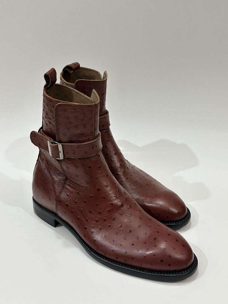 May include: A pair of brown ankle boots featuring a textured ostrich-style leather finish with a distinct quill pattern. Each boot includes a side strap with a silver-toned metal buckle, a pull tab at the heel, and a solid black sole.