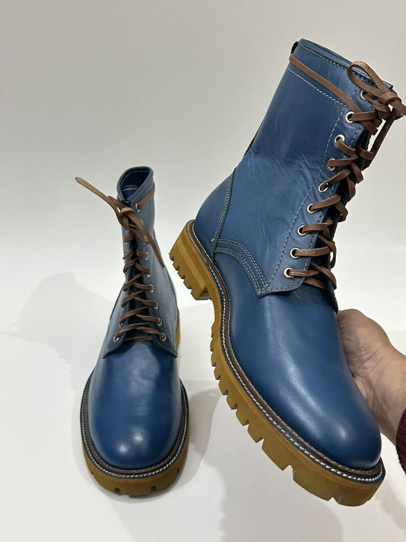 以下が含まれることがあります： 茶色の靴紐、銀色の金属製アイレット、厚みのあるベージュのラバーラグソールを備えた青いレザーのレースアップブーツ。ブーツは滑らかな質感で、ウェルトとアッパーパネルに沿って目に見えるステッチが施されています。