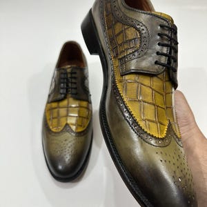 Puede incluir: Un par de zapatos oxford de cuero con un esquema de color bicolor en verde oliva y amarillo mostaza. Los zapatos tienen un patr&oacute;n texturizado con relieve de cocodrilo en el empeine y el lateral, cordones negros y detalles brogue.
