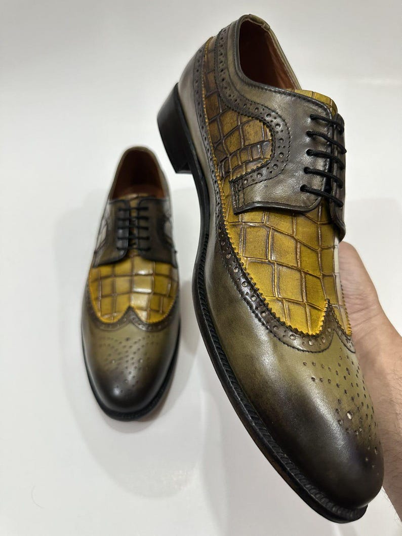 Puede incluir: Un par de zapatos oxford de cuero con un esquema de color bicolor en verde oliva y amarillo mostaza. Los zapatos tienen un patr&oacute;n texturizado con relieve de cocodrilo en el empeine y el lateral, cordones negros y detalles brogue.