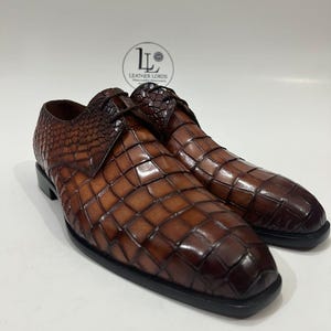 May include: A pair of brown leather dress shoes featuring a textured crocodile-skin pattern with a gradient finish, dark brown laces, and a black sole. A circular logo with the text LEATHER LORDS is visible in the background.