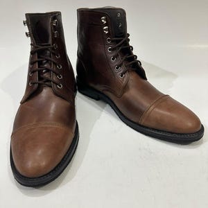 May include: A pair of brown leather lace-up boots featuring a cap toe design, dark brown laces, and silver-toned metal eyelets and speed hooks. The boots have a smooth leather finish and a thick black rubber sole with a defined heel.