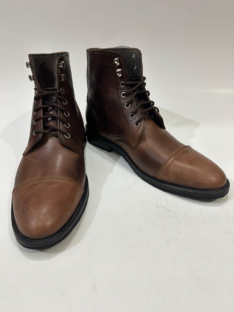 May include: A pair of brown leather lace-up boots featuring a cap toe design, dark brown laces, and silver-toned metal eyelets and speed hooks. The boots have a smooth leather finish and a thick black rubber sole with a defined heel.