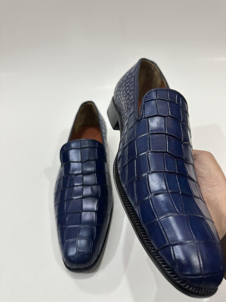 May include: A pair of navy blue loafers featuring a textured crocodile-embossed pattern across the upper. The slip-on shoes have a rounded toe, a low heel, and a smooth tan interior lining, showcasing a structured and polished appearance.