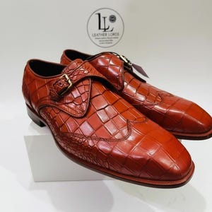 May include: A pair of cognac-colored monk strap shoes featuring a textured crocodile-embossed pattern, a gold-toned metal buckle, and a wingtip design. The logo for Leather Lords is visible in the background above the footwear.
