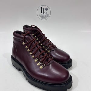 May include: A pair of burgundy leather ankle boots with dark red laces, brass-toned metal eyelets, and a thick black lug sole. The boots feature a logo on the upper heel area with the text LEATHER LORDS Where Leather Meets Luxury.
