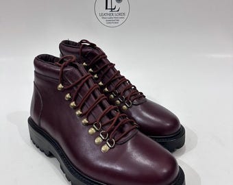 May include: A pair of burgundy leather ankle boots featuring a thick black lug sole, matching burgundy laces, and gold-toned metal D-ring eyelets. A circular logo above the boots displays the text LEATHER LORDS Where Leather Meets Luxury.