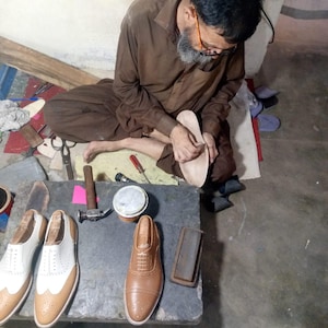 May include: A pair of white and tan leather wingtip oxford shoes and a single tan leather oxford shoe on a stone workbench. The shoes feature intricate brogue detailing, lace-up closures, and wooden shoe trees inserted inside the footwear.