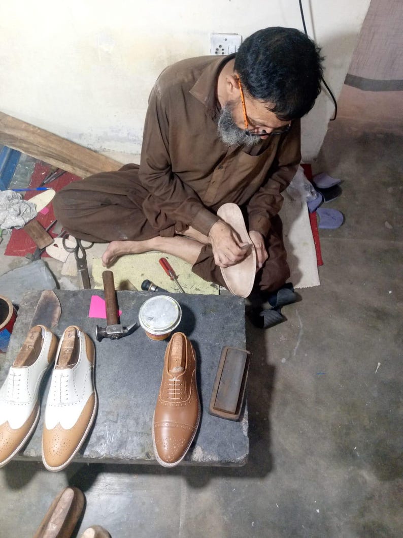 May include: A pair of white and tan leather wingtip oxford shoes and a single tan leather oxford shoe on a stone workbench. The shoes feature intricate brogue detailing, lace-up closures, and wooden shoe trees inserted inside the footwear.