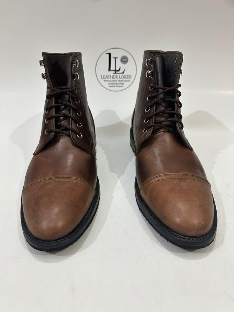 May include: A pair of brown leather lace-up boots with cap toes, silver-toned metal eyelets, and black soles. A circular logo with the text LEATHER LORDS is positioned between the boots, featuring a stylized LL monogram and text.