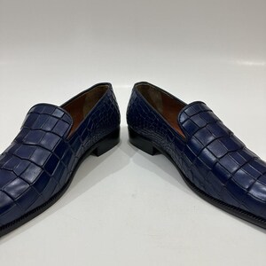 May include: A pair of deep blue loafers featuring a textured crocodile-embossed leather upper, a low black heel, and a smooth brown interior lining. The slip-on shoes have a structured silhouette with a rounded toe and visible stitching.