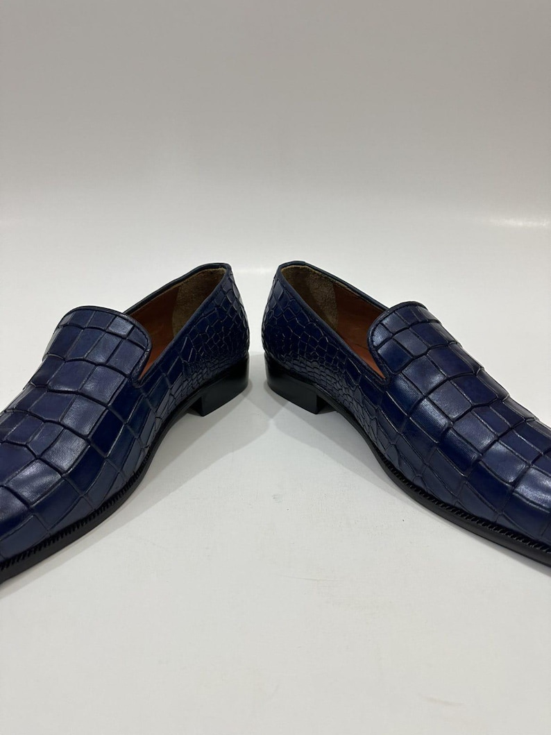 May include: A pair of deep blue loafers featuring a textured crocodile-embossed leather upper, a low black heel, and a smooth brown interior lining. The slip-on shoes have a structured silhouette with a rounded toe and visible stitching.