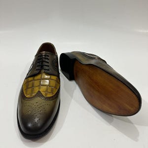 Puede incluir: Un par de zapatos oxford con punta de ala que presentan un acabado en cuero degradado verde oliva y marr&oacute;n. El empeine muestra un patr&oacute;n texturizado con relieve de cocodrilo en amarillo mostaza, cordones negros y detalles brogue.