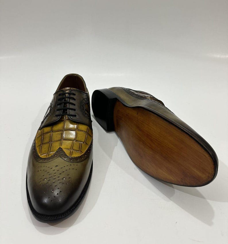 Puede incluir: Un par de zapatos oxford con punta de ala que presentan un acabado en cuero degradado verde oliva y marr&oacute;n. El empeine muestra un patr&oacute;n texturizado con relieve de cocodrilo en amarillo mostaza, cordones negros y detalles brogue.