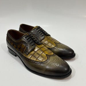 May include: A pair of olive green and brown leather oxford shoes featuring a wingtip design with brogue detailing and a textured crocodile-embossed pattern on the vamp and side panels. The shoes include black laces and a smooth leather finish.