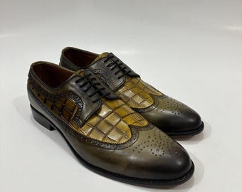 May include: A pair of olive green and brown leather oxford shoes featuring a wingtip design with brogue detailing and a textured crocodile-embossed pattern on the vamp and side panels. The shoes include black laces and a smooth leather finish.
