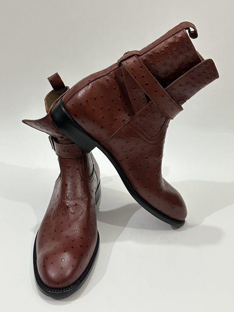May include: A pair of brown ankle boots featuring a textured ostrich-style leather finish with a distinct quill pattern. Each boot includes a wrap-around strap with a metal buckle, a pull tab at the heel, and a solid black sole and heel.