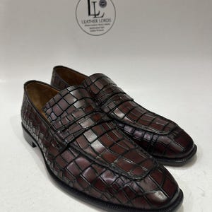 Puede incluir: Un par de mocasines de cuero marrón con un patrón texturizado similar al de un cocodrilo. Los zapatos tienen una puntera cuadrada y una correa en la parte superior. El logotipo de la marca "Leather Lords" es visible en el fondo.