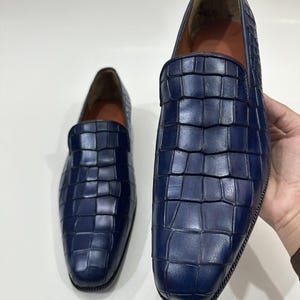 May include: A pair of deep blue loafers featuring a textured crocodile-embossed pattern across the upper surface. The slip-on shoes have a rounded toe, a smooth interior lining, and a dark, ridged sole edge visible along the bottom perimeter.