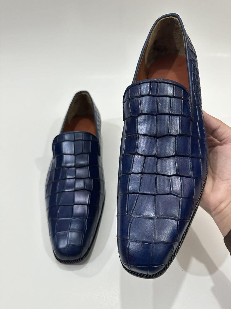 May include: A pair of deep blue loafers featuring a textured crocodile-embossed pattern across the upper surface. The slip-on shoes have a rounded toe, a smooth interior lining, and a dark, ridged sole edge visible along the bottom perimeter.